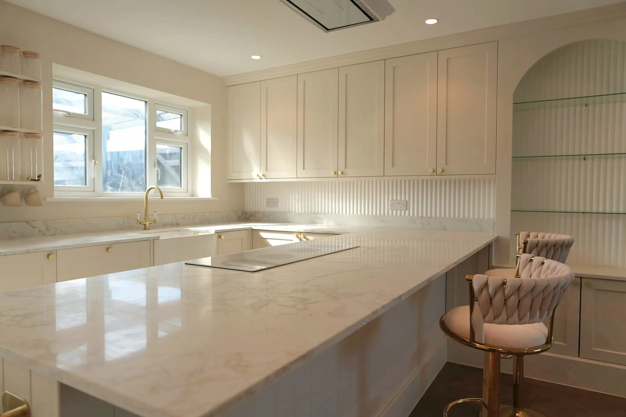 A bright cream shaker kitchen with a marble-effect worktop, fluted splashback and brass bar stools