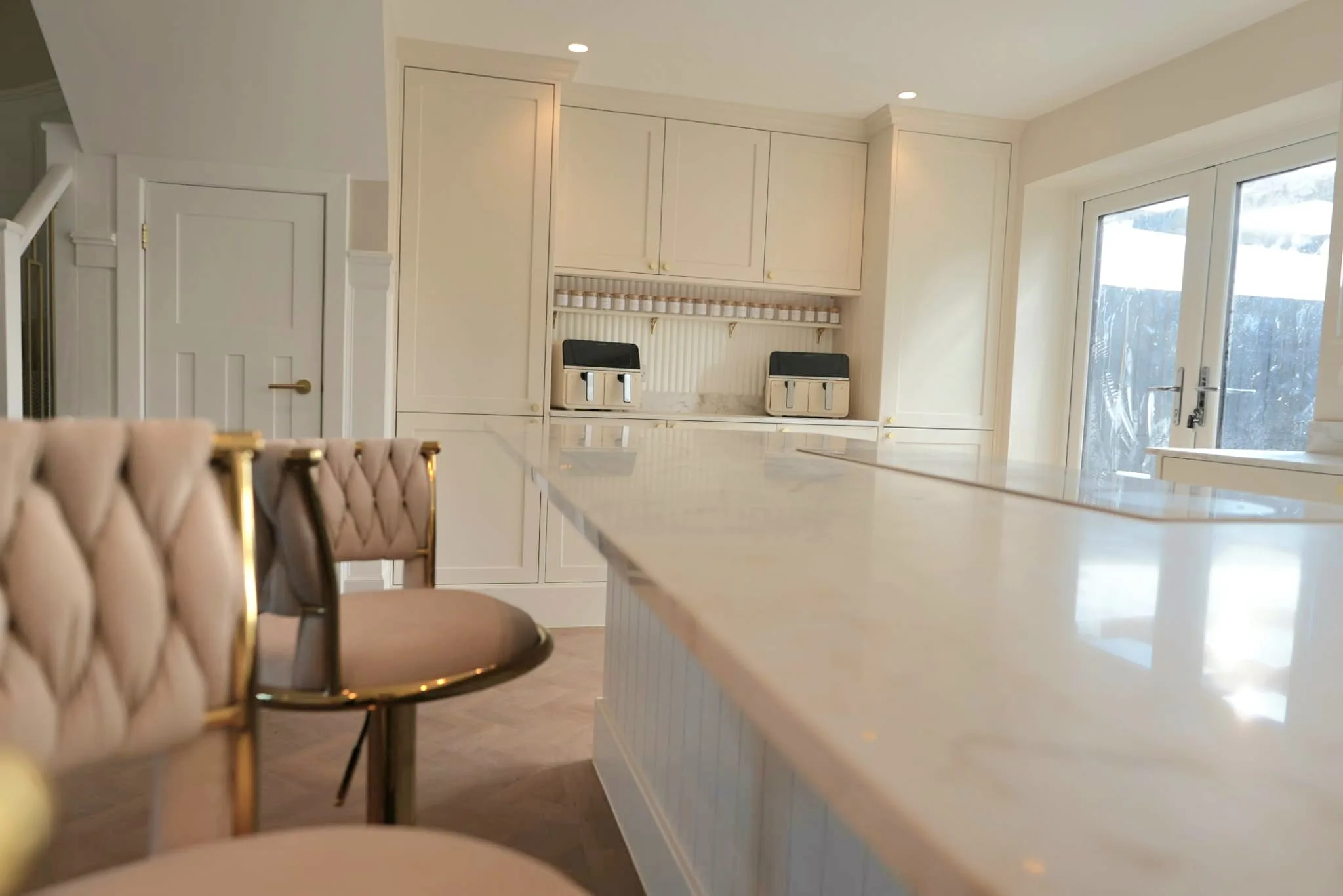 A cream shaker kitchen island with a marble-effect worktop, brass bar stools and tall pantry cabinets